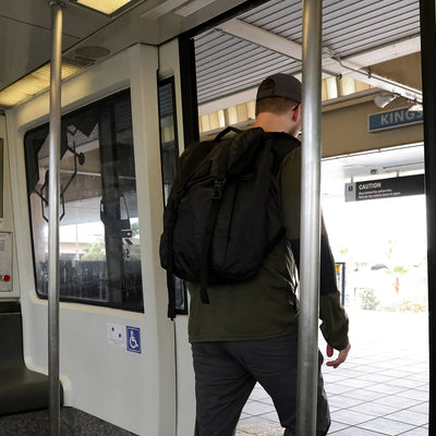 A man wearing a long-sleeved green shirt and gray pants exits a train at the station, with a GORUCK M23 - Dyneema bag resembling military gear on his shoulder. The train doors stay open, and the station name sign is partially visible in the background.