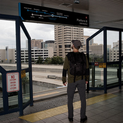 A person sporting a GORUCK M23 - Dyneema rucksack and cap stands on a train station platform, gazing at an electronic sign listing upcoming stations. In the background, tall buildings form a cityscape. A No Loitering sign is displayed on a window.