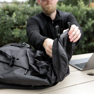 A person is adjusting the strap of a GORUCK M23 - Dyneema backpack on an outdoor table. A laptop is partially visible next to them, and the background showcases greenery.