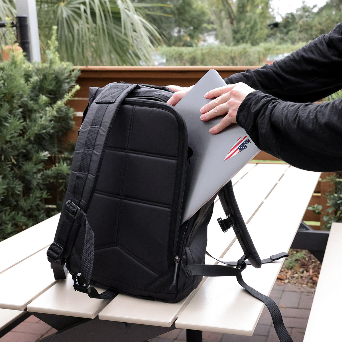 A person in a black long-sleeve shirt places a laptop with an American flag sticker into the sleek, military-inspired GORUCK M23 - Dyneema bag on a picnic table. The background showcases greenery and a wooden fence.