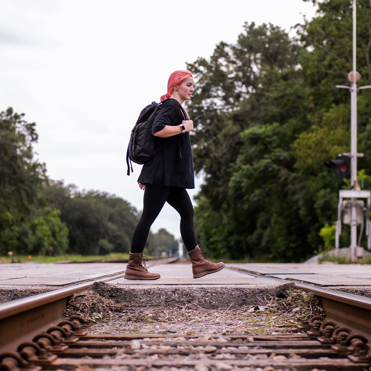 A person with pink hair confidently crosses railroad tracks, surrounded by trees and a cloudy sky, wearing a GORUCK M23 - Dyneema® backpack. They are dressed in a black shirt, leggings, and brown boots.