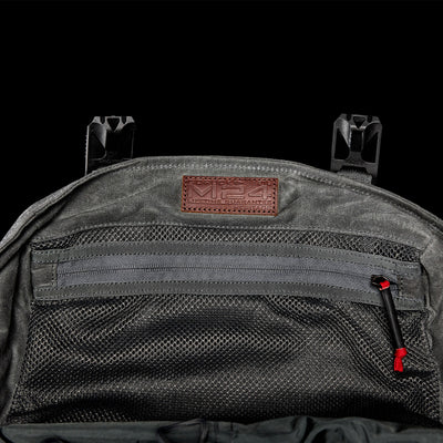 Here's a revised version of the sentence using the provided product data:

Close-up of a waxed canvas backpack in gray with a mesh zippered pocket, crafted from durable materials by GORUCK. The bag features a brown leather patch embossed with M24. Two black clips are visible at the top against a solid dark background, adding to its modern rucksack appeal.