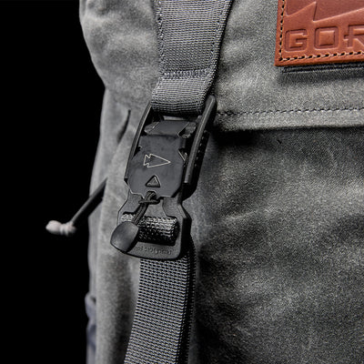 Close-up of a gray M24 - Waxed Canvas rucksack's buckle and strap, featuring a secure plastic clasp and the durable materials' subtle textures. A brown leather patch with partial text from GORUCK is visible on the top right, enhancing the modern rucksack's sleek design.