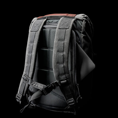 A sleek, black M24 Waxed Canvas rucksack from GORUCK with padded, adjustable shoulder straps is shown against a black background. A laptop peeks out from a partially open side pocket, highlighting the bag's functionality and durable materials that enhance its storage capabilities.