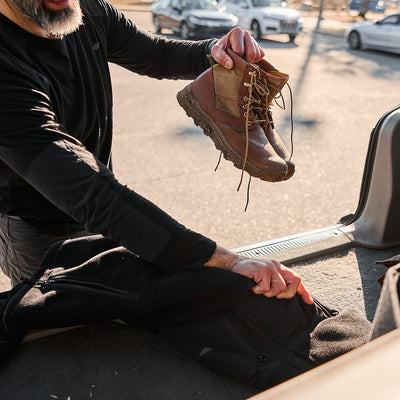 A person carefully places a MACV-2 - Mid Top - Briar + Coyote with an aggressive triple compound outsole into a black bag in the back of a vehicle, surrounded by parked cars.