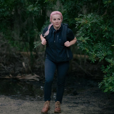 A person with pink hair hikes a wooded trail, wearing a backpack and boots with the MACV-2 - Mid Top in Briar + Coyote, boasting an aggressive triple compound outsole for superior grip.