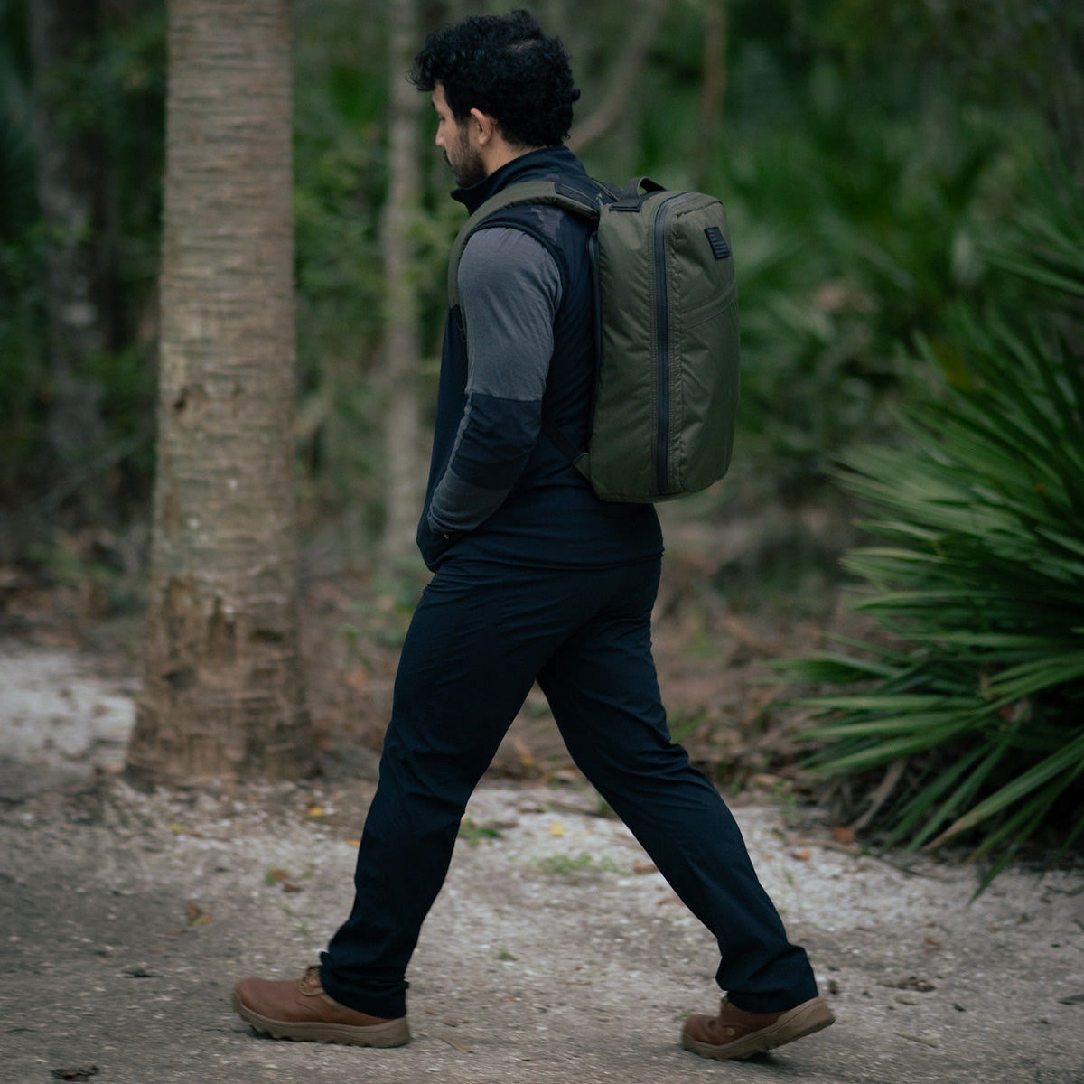 A person wearing MACV-2 - Mid Top - Briar + Coyote backpack walks on a forest path, their jungle boots gripping the terrain with an aggressive triple compound outsole, surrounded by lush green foliage.