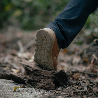 The MACV-2 - Mid Top - Briar + Coyote boot steps over a log on a forest trail, its aggressive triple compound outsole gripping the earth amidst leaves and dirt.