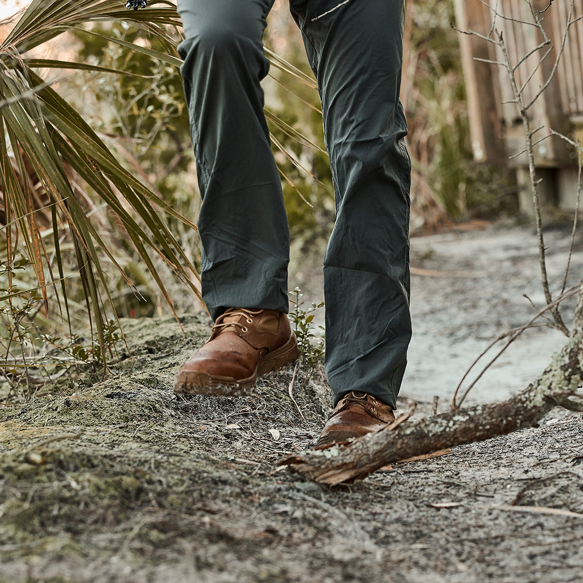 A person hiking on a forest trail, wearing green pants and sturdy MACV-2 Mid Top boots in Briar + Coyote, featuring an aggressive triple compound outsole for maximum traction.