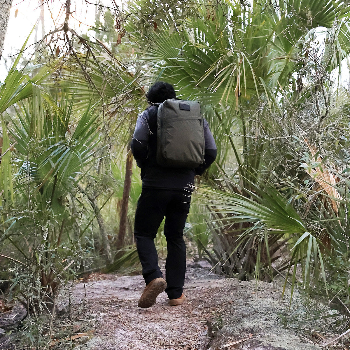 A person hiking on a dirt path through dense greenery with a large backpack, confidently sporting MACV-2 - Mid Top - Briar + Coyote boots equipped with an aggressive triple compound outsole.