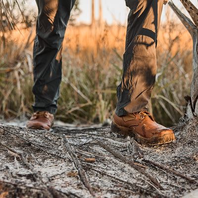 A person wearing MACV-2 jungle boots with an aggressive triple compound outsole and gray pants walks along a muddy path, surrounded by dry grass.