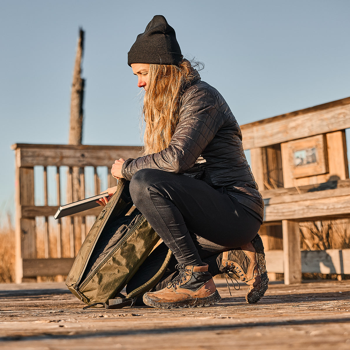 A woman in outdoor gear methodically packs her MACV-2 Mid Top in Chestnut + Black and other essentials into a backpack on a wooden platform under clear skies.