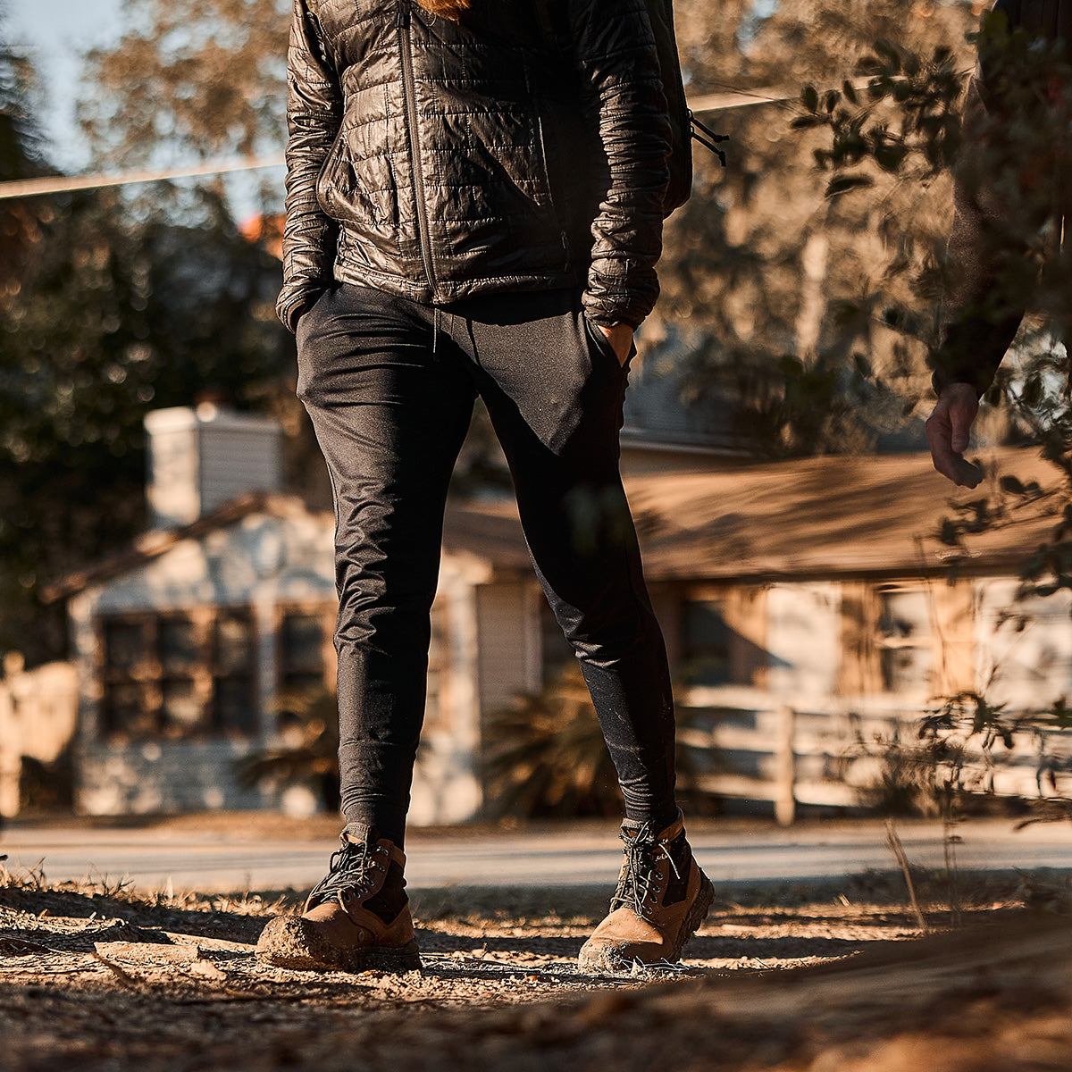 Strolling on a sunny path in a black jacket and pants, a person sports the MACV-2 - Mid Top - Chestnut + Black boots with a triple compound outsole, while the house and trees stand quietly behind them.