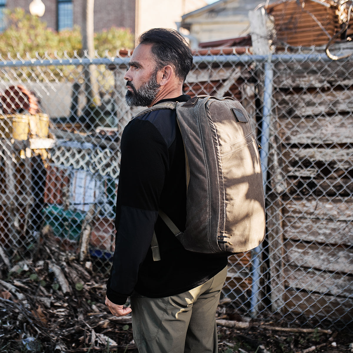 A man adorned in the Men’s Commando Long Sleeve - Merino Wool by GORUCK and carrying a backpack is positioned near a fence with stacked wooden pallets in the backdrop.