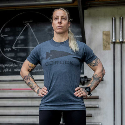 A person with long hair in a ponytail and tattoos on both arms stands confidently in a gym. Wearing the GORUCK Spearhead Tee - Tri-Blend from vendor-unknown and black pants, they embody the Special Forces roots. Behind them, barbells and a blackboard with chalk drawings enhance the rugged atmosphere.