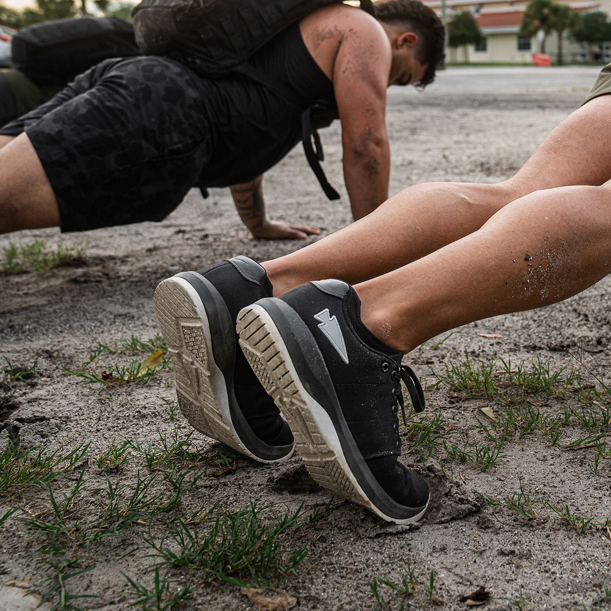 Two individuals are performing outdoor push-ups in a sandy, gritty environment, each entirely focused. One of them is wearing the Men's Ballistic Trainers by GORUCK in Black and Glacier Grey with a Silver Reflective Spearhead, designed for optimal functional fitness performance. Surrounded by small patches of grass, their determination is evident.