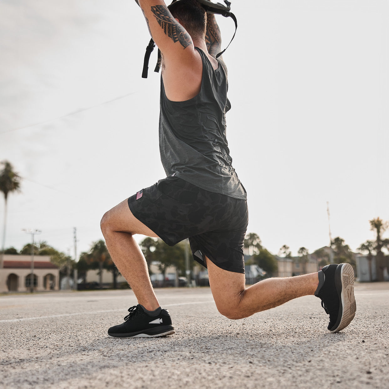 Wearing GORUCK's Men's Ballistic Trainers in Black + Glacier Grey with a Silver Reflective Spearhead, a person performs a lunge while holding a kettlebell overhead. They exercise outdoors on a sunny day, with palm trees and buildings in the background, highlighting their functional fitness footwear.