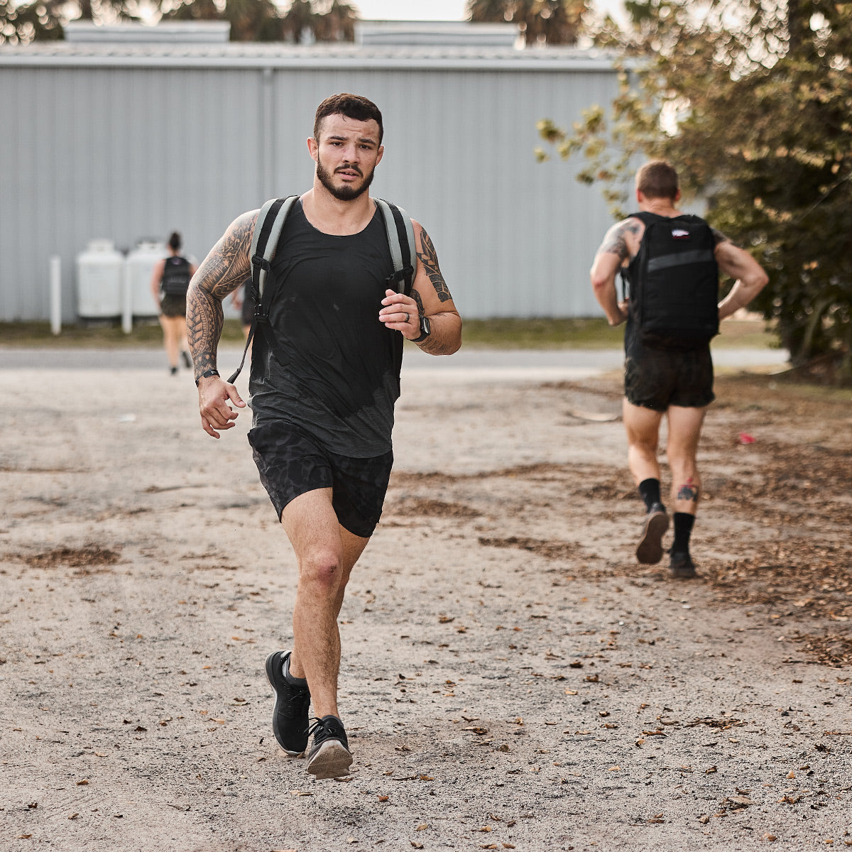 A man with tattoos and a backpack sprints down the dirt path in his GORUCK Men's Ballistic Trainers - Black + Glacier Grey with Silver Reflective Spearhead, sporting a black tank top and shorts. Another man runs ahead in a similar outfit. Trees and a building form the backdrop of their energetic run.