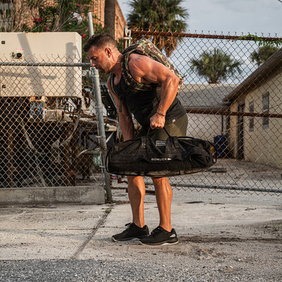 A man in athletic wear hoists a weighted black sandbag outdoors, wearing GORUCK Men's Ballistic Trainers in Black and Glacier Grey with a Silver Reflective Spearhead. He sports a camouflage backpack made from CORDURA® Ballistic Nylon. Positioned on a rough concrete surface with a chain-link fence, utility boxes, and palm trees behind him, he exemplifies peak functional fitness.