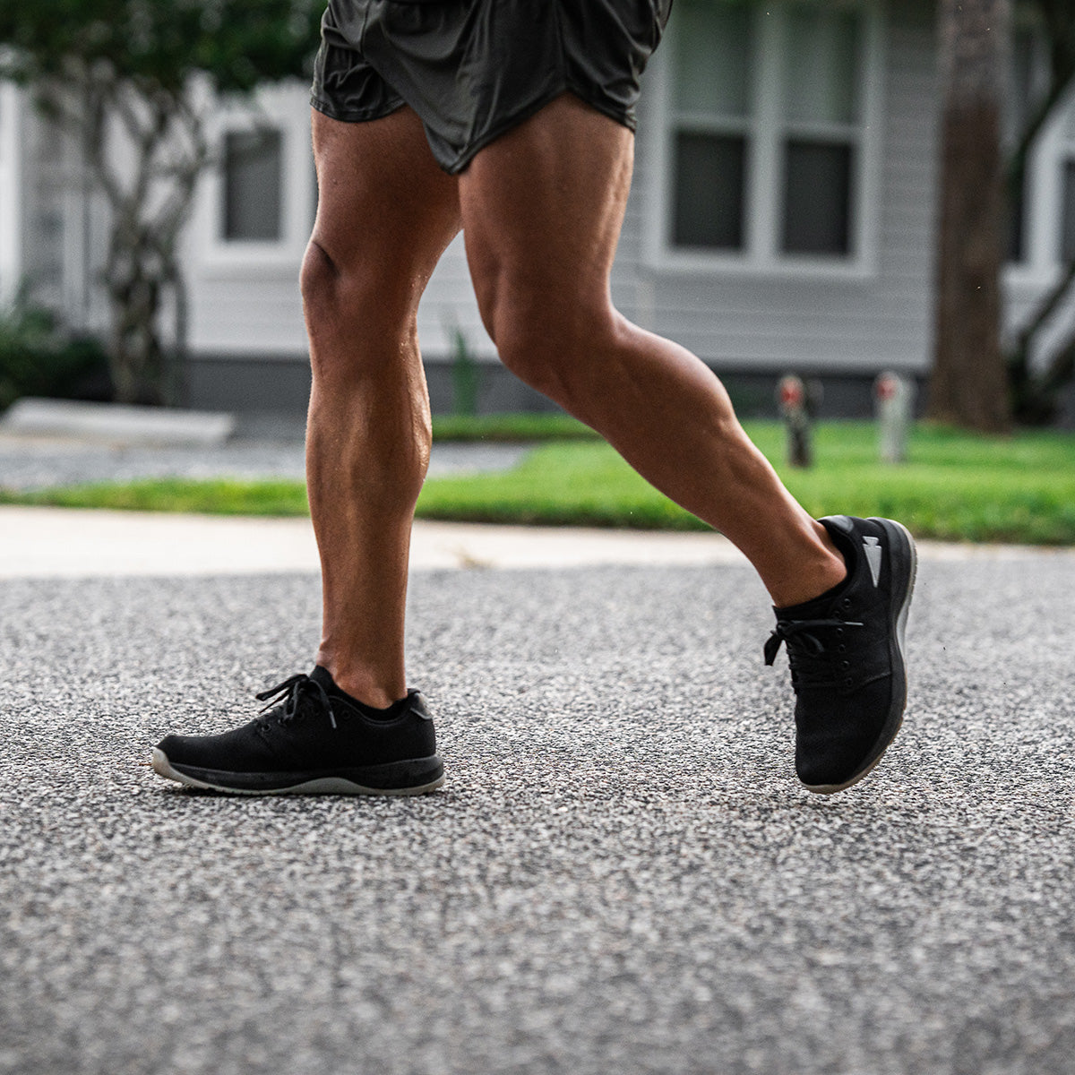 A person in black GORUCK Men's Ballistic Trainers - Black + Glacier Grey W / Silver Reflective Spearhead and shorts is jogging on a street. The background features trees and a house with white windows. The sunlight highlights the person's toned legs and the textured road surface, showcasing their functional fitness footwear.