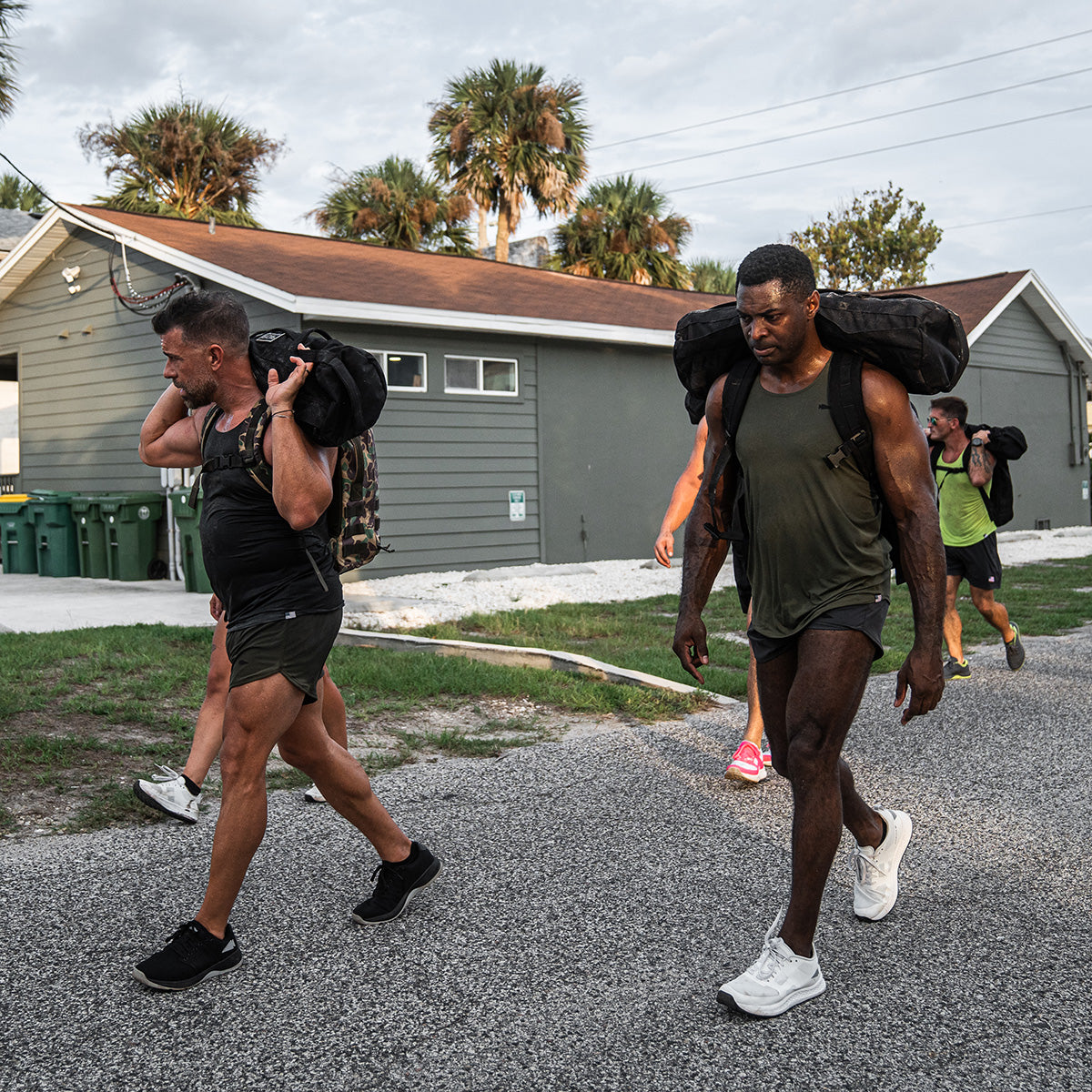 A group of people walk along a road carrying heavy sandbags on their shoulders, clad in GORUCK Men's Ballistic Trainers in Black and Glacier Grey with a Silver Reflective Spearhead, perfect for such challenges. In the background, palm trees sway near a house with a brown roof.