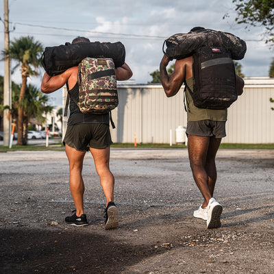 Two individuals are walking on gravel, each shouldering a large duffel bag. One has a camouflage backpack crafted from CORDURA® Ballistic Nylon, while the other carries a black backpack. Both are sporting GORUCK's Men's Ballistic Trainers in Black and Glacier Grey with Silver Reflective Spearhead, paired with athletic shorts and tank tops as they head toward a building.