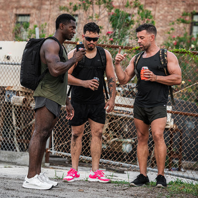 Three men dressed in athletic gear, with GORUCK backpacks and Men's Ballistic Trainers in Black and Glacier Grey featuring a Silver Reflective Spearhead design, stand near a chain-link fence. They are holding drinks and seem to be engaged in conversation, with trees and a brick building visible in the background.