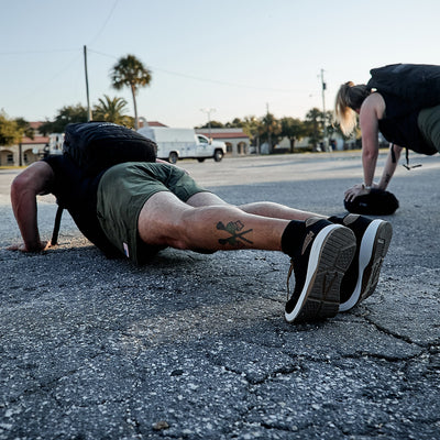 Two individuals are doing push-ups on an outdoor asphalt surface, highlighting their functional fitness footwear and athletic attire. Each person carries a backpack made from durable CORDURA Ballistic Nylon. A palm tree and buildings are visible in the background, as the black and white Men's Ballistic Trainers by GORUCK with the Coyote Reflective Spearhead design draw attention to the foreground figure with a leg tattoo.