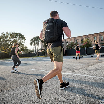 A group of people in athletic wear runs outdoors in a parking lot during the day. Prominent in the foreground is a man wearing Men's Ballistic Trainers by GORUCK in Black + White with Coyote Reflective Spearhead and carrying a black backpack made from CORDURA Ballistic Nylon. Trees and a building provide the backdrop for this display of functional fitness.
