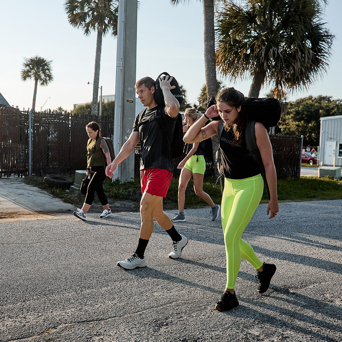 Under the sun, a group of four people strides effortlessly, each carrying weighted bags. Dressed in athletic wear and GORUCK's Men's Ballistic Trainers in Lunar Rock and Charcoal with Silver Reflective Spearhead, they enjoy the scenic backdrop of palm trees. These trainers provide 3X Stability, enabling them to showcase their strength and endurance.