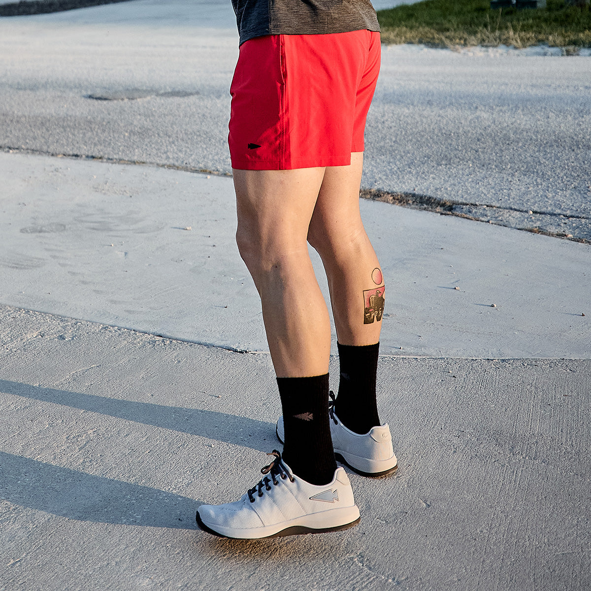 A person is standing on a paved surface, dressed in a gray top and vibrant red shorts, with black socks and GORUCK's Men's Ballistic Trainers in Lunar Rock and Charcoal featuring a Silver Reflective Spearhead design. A tattoo adorns the lower left leg against a backdrop of a road bordered by grassy areas.