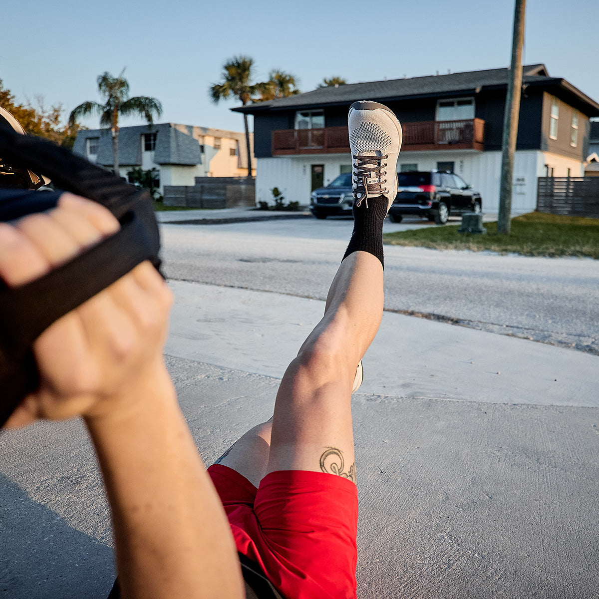 Exercising outdoors by lying on the ground, a person lifts their right leg straight up while wearing black socks and light-colored Men's Ballistic Trainers from GORUCK, featuring the 3X Stability system. They are also dressed in red shorts and holding a fitness band, with residential homes providing the backdrop for this showcase of functional fitness footwear expertise.