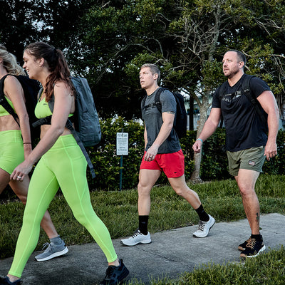 Four individuals walk in line on the sidewalk, highlighting their athletic clothing and GORUCK footwear. The person at the front is dressed in a vivid green ensemble, while the others choose darker tops paired with shorts. Each of them carries a backpack that might be made from durable CORDURA® Ballistic Nylon. Lush trees serve as a scenic backdrop for those wearing Men's Ballistic Trainers in Lunar Rock and Charcoal with Silver Reflective Spearhead accents.