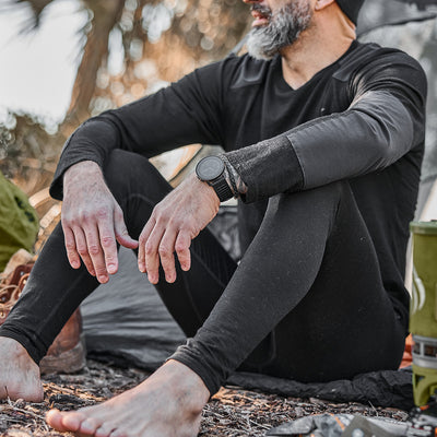 A man in black outdoor attire sits on the ground among trees, confident and comfortable with his breathable gear. Wearing Men's Base Layer Bottom - Merino Wool, backed by Scars Lifetime Guarantee, he embraces his adventure.