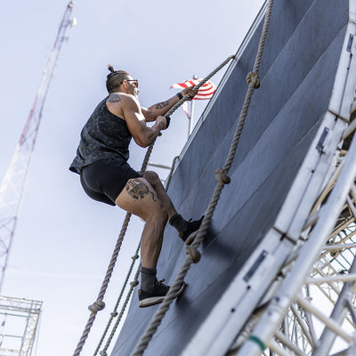 Wearing the Men’s Ranger Panties from GORUCK, crafted with ToughStretch fabric, a man scales a steep obstacle wall using ropes. His athletic attire shines in the sunlight, revealing tattoos on his arms and legs, as the American flag flutters proudly against the clear sky.