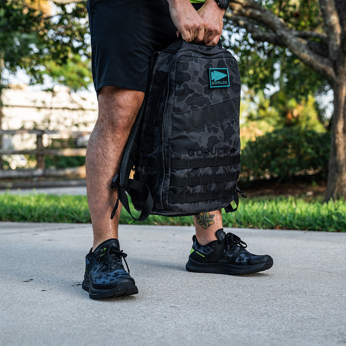 A person in black shorts and sneakers, equipped with a GORUCK Men's Rough Runner - Midnight Frogskin + Acid Lime backpack, stands on a paved path. The scene features green grass and a tree in the background, blending the rugged spirit of adventure with serene nature.