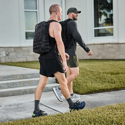 Two men walking on a sidewalk next to a grassy area, dressed in athletic gear with GORUCK Rough Runner backpacks in Midnight Frogskin and Acid Lime. One of the men wears a cap as they pass by a building with large windows in the background.