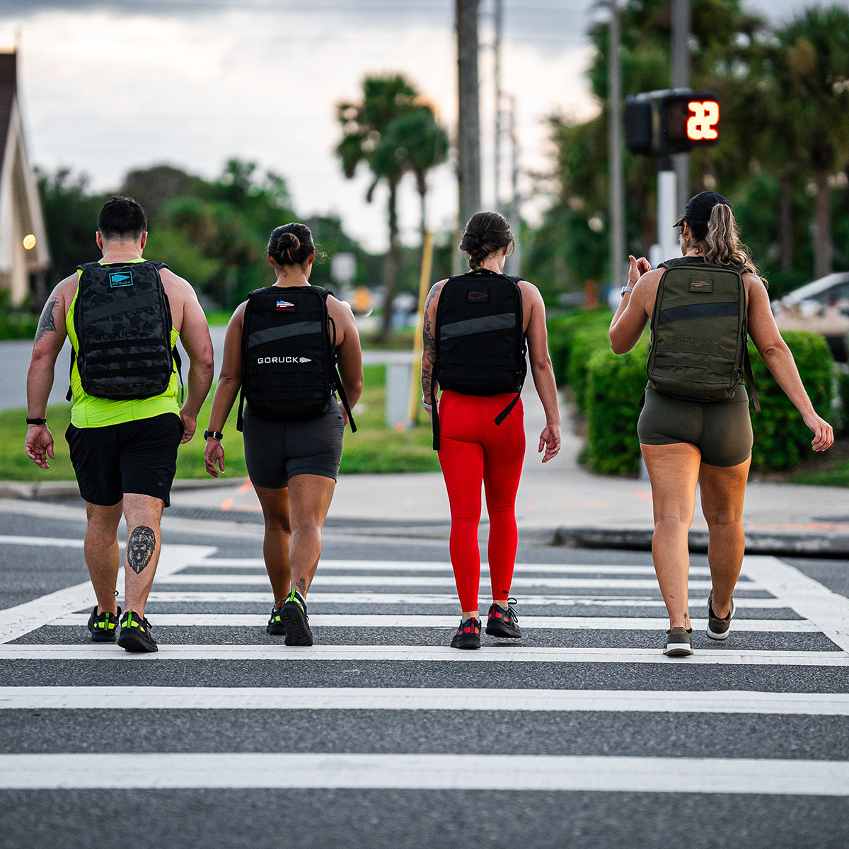 Four individuals equipped with Men's Rough Runner - Midnight Frogskin + Acid Lime backpacks from GORUCK make their way across the street at a crosswalk. The illuminated walk signal guides them under the sun, as palm trees sway alongside contemporary architecture, embodying an active spirit and hinting at the energetic essence of Rough Runner™ in this dynamic outdoor scene.