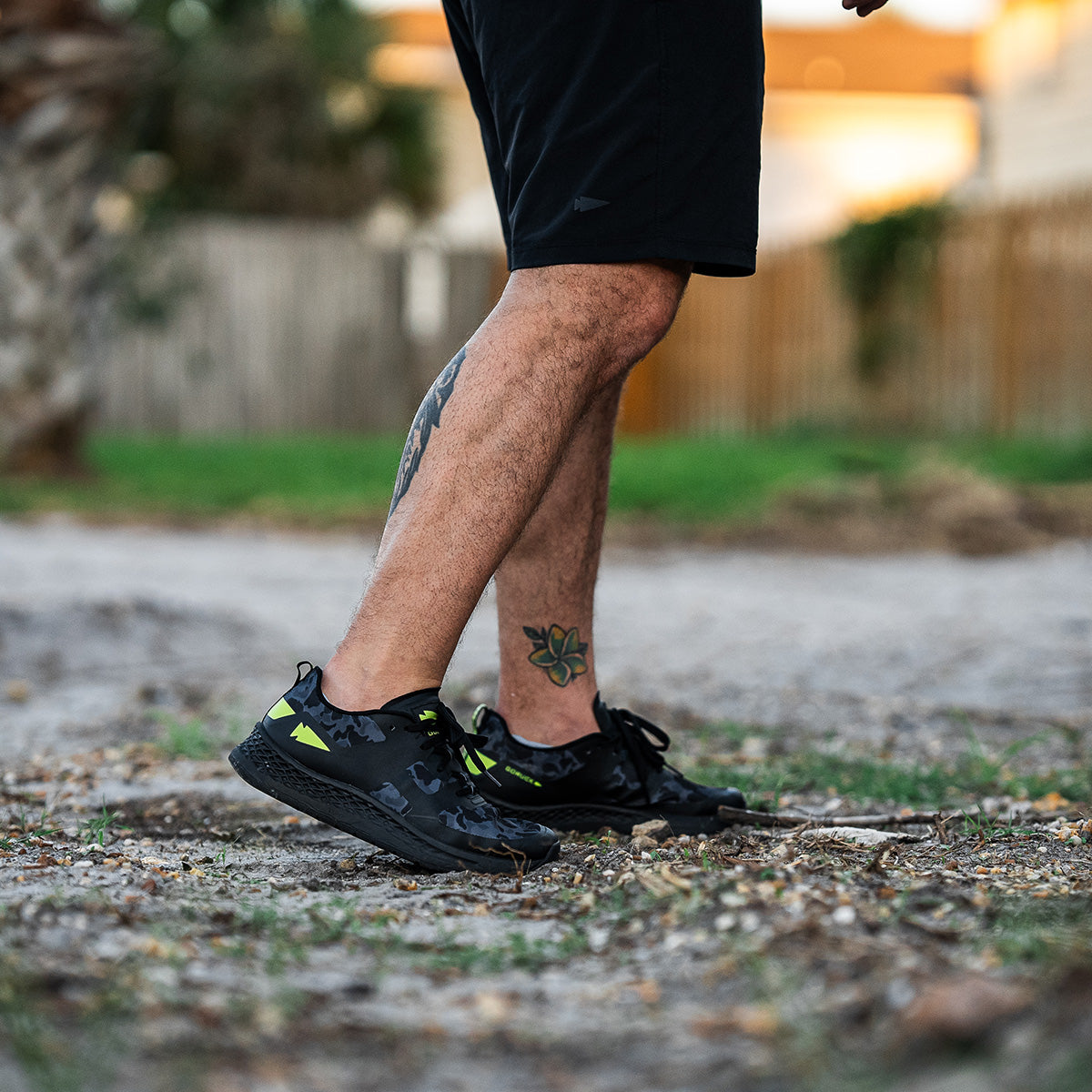 A person donning black shorts and GORUCK's Men's Rough Runner - Midnight Frogskin + Acid Lime sneakers strides along a dirt path. Their tattooed legs add character to the scene, framed by a wooden fence and out-of-focus greenery, creating the perfect backdrop for this energetic walk.