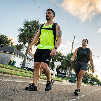 As the sun sets, two individuals walk briskly down a suburban street, outfitted in athletic gear and carrying backpacks from Rough Runner™. The man is dressed in a neon tank top and shorts from GORUCK's Men's Rough Runner collection in Midnight Frogskin + Acid Lime, while the woman opts for a black tank top and shorts. The scene is framed by palm trees and houses, creating a picturesque view.