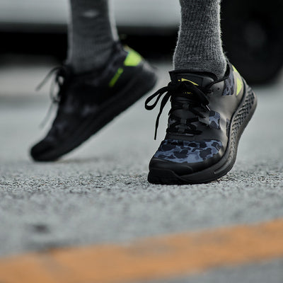 Close-up of a person wearing the Men's Rough Runner - Midnight Frogskin + Acid Lime by GORUCK, showcasing black and green athletic shoes with a camouflage pattern and EVA Midsole, running on a textured outdoor surface. The focus is on the shoes, with legs in gray socks visible.