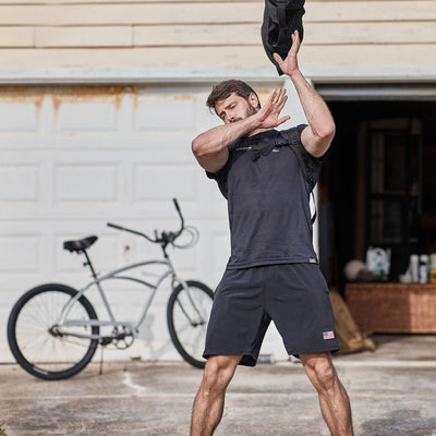 A man wearing a black GORUCK ToughStretch t-shirt and Men’s USA Training Shorts is outside, swinging a black bag over his shoulders. A bicycle stands parked against a white garage door in the background—a setting designed to endure, just like the SCARS Lifetime Guarantee.