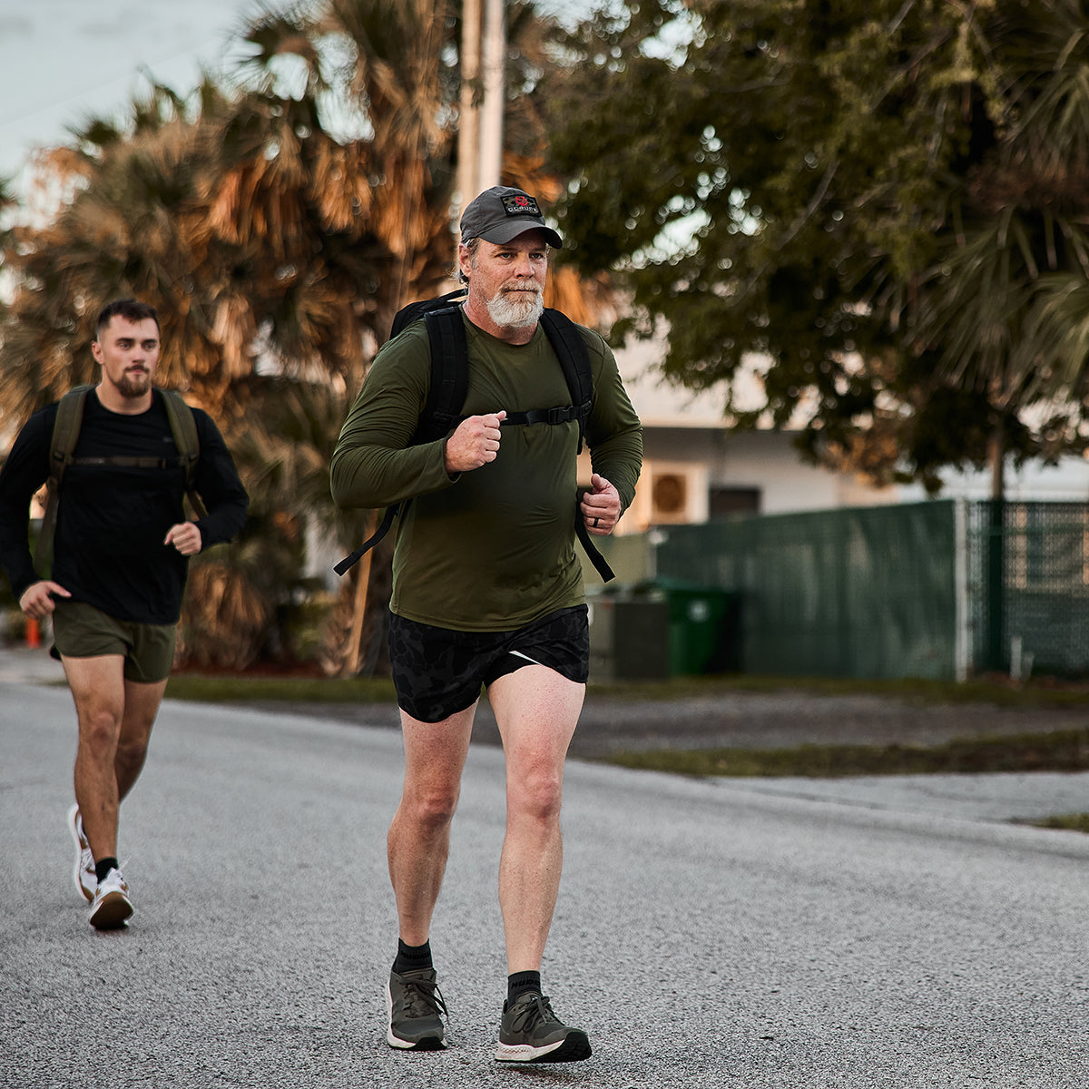 Two men walk briskly on a residential street, dressed in casual athletic gear and carrying backpacks. The older man leads in olive green while the younger follows, sporting GORUCK Men’s Ranger Panties made with ToughStretch fabric. Trees and a house stand quietly in the background.