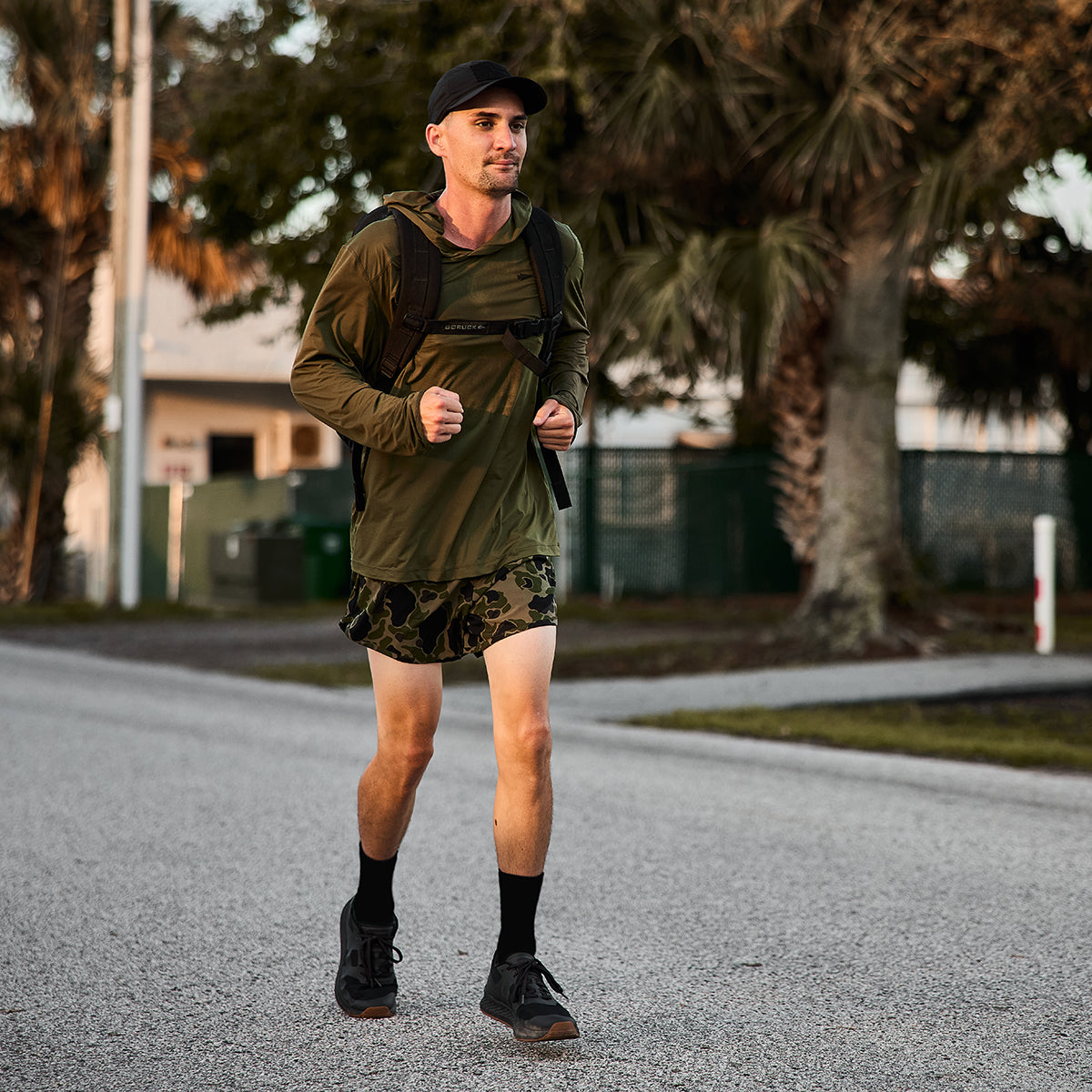 A man briskly walks on a suburban street, wearing a green long-sleeve shirt, Men’s Ranger Panties - ToughStretch by GORUCK, a black cap, and carrying a backpack. He is surrounded by trees and houses in the background.