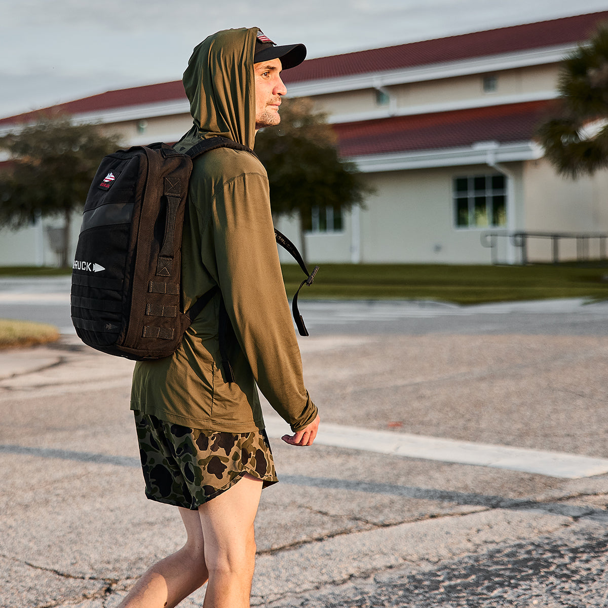 A person sporting an olive green hoodie and GORUCK’s camo Men's Ranger Panties - ToughStretch shorts is spotted outdoors with a black backpack. The background features a building with a red roof, trees, and a clear sky. The ToughStretch fabric of their attire guarantees comfort as they explore the scenery.