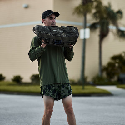 A person wearing a green shirt and GORUCK Men’s Ranger Panties - ToughStretch shorts is carrying a camo sandbag on their shoulders outdoors. With a black cap, they confidently stand on a paved area surrounded by palm trees and a building, assured by the Scars Lifetime Guarantee.