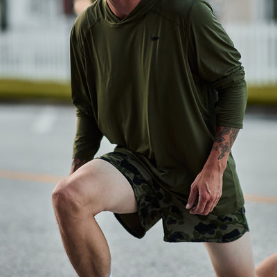 A person in a loose dark green hoodie is stretching outdoors, preparing for exercise, wearing GORUCK's Men’s Ranger Panties made from ToughStretch fabric. The background shows a street and a white fence. Tattoos are visible on the person's arm as their camouflage shorts provide maximum flexibility.