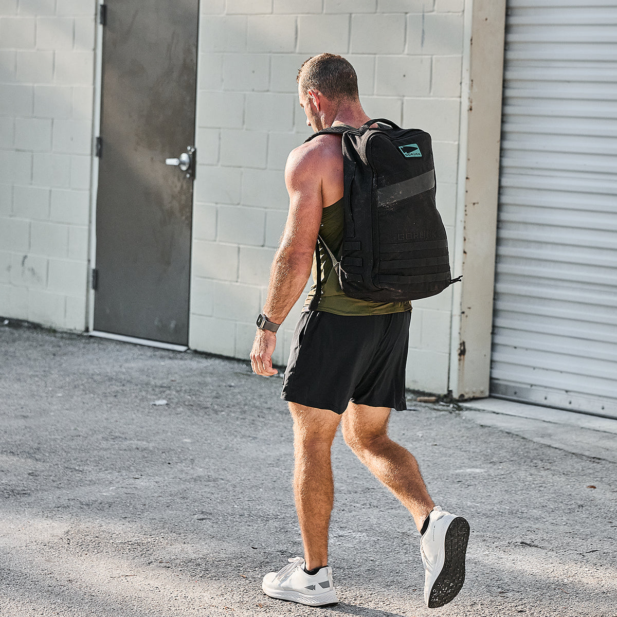 A man outfitted in athletic apparel, featuring GORUCK's Men’s USA Training Shorts made with ToughStretch material, carries a large black backpack. He walks confidently on a concrete surface past a white brick wall that has a door and closed garage door. The area is illuminated by bright sunlight.