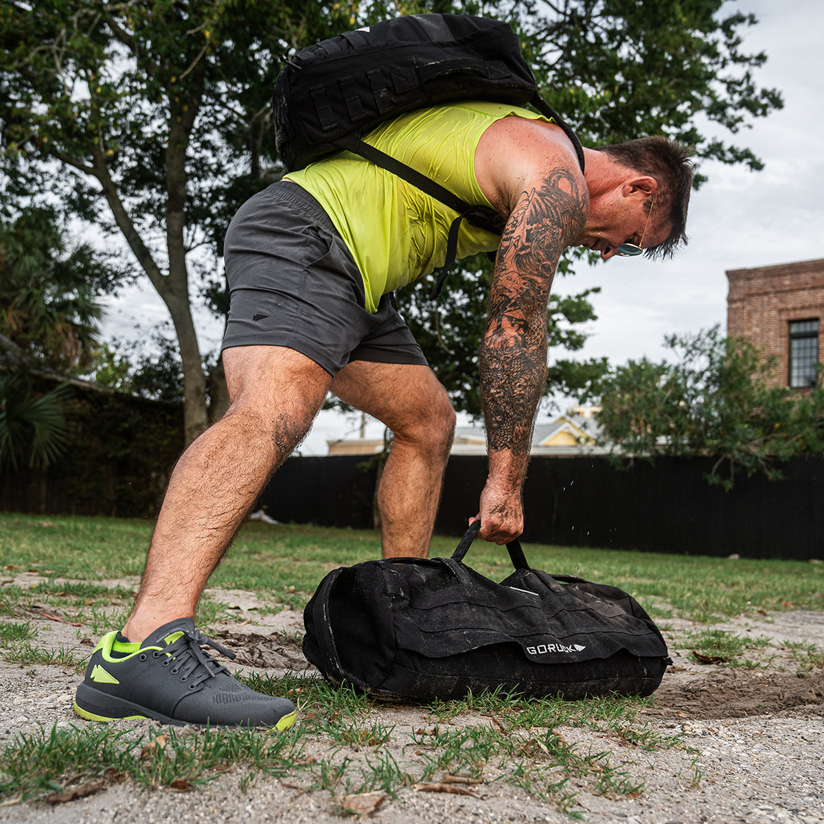 A person wearing a yellow tank top and GORUCK’s Men’s USA Training Shorts - ToughStretch bends forward, lifting a black sandbag outdoors. They have a tattooed arm, green-accented sneakers, and a backpack. Trees and a brick building stand in the background.