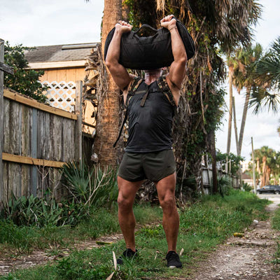 Clad in GORUCK's Men’s Ranger Panties made from ToughStretch fabric and a tank top, a person lifts a heavy sandbag overhead outdoors. The scene is set against a backdrop of rustic fencing, palm trees, and vibrant greenery along the pathway. These shorts are designed to provide ultimate comfort and performance for such physically demanding tasks.